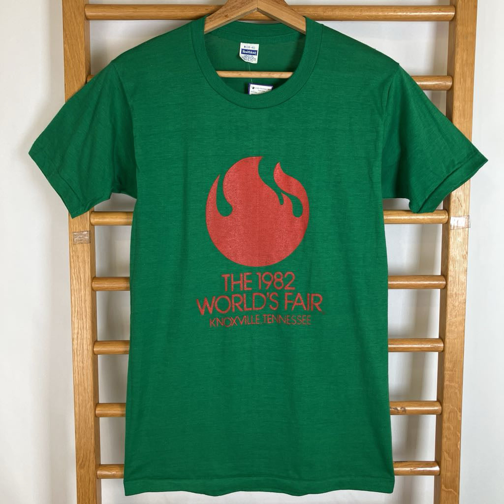 A green short-sleeve t-shirt hanging on a wooden hanger against a wooden ladder. The shirt features a red flame graphic and the text 'THE 1982 WORLD'S FAIR' and 'KNOXVILLE TENNESSEE' printed on the front.