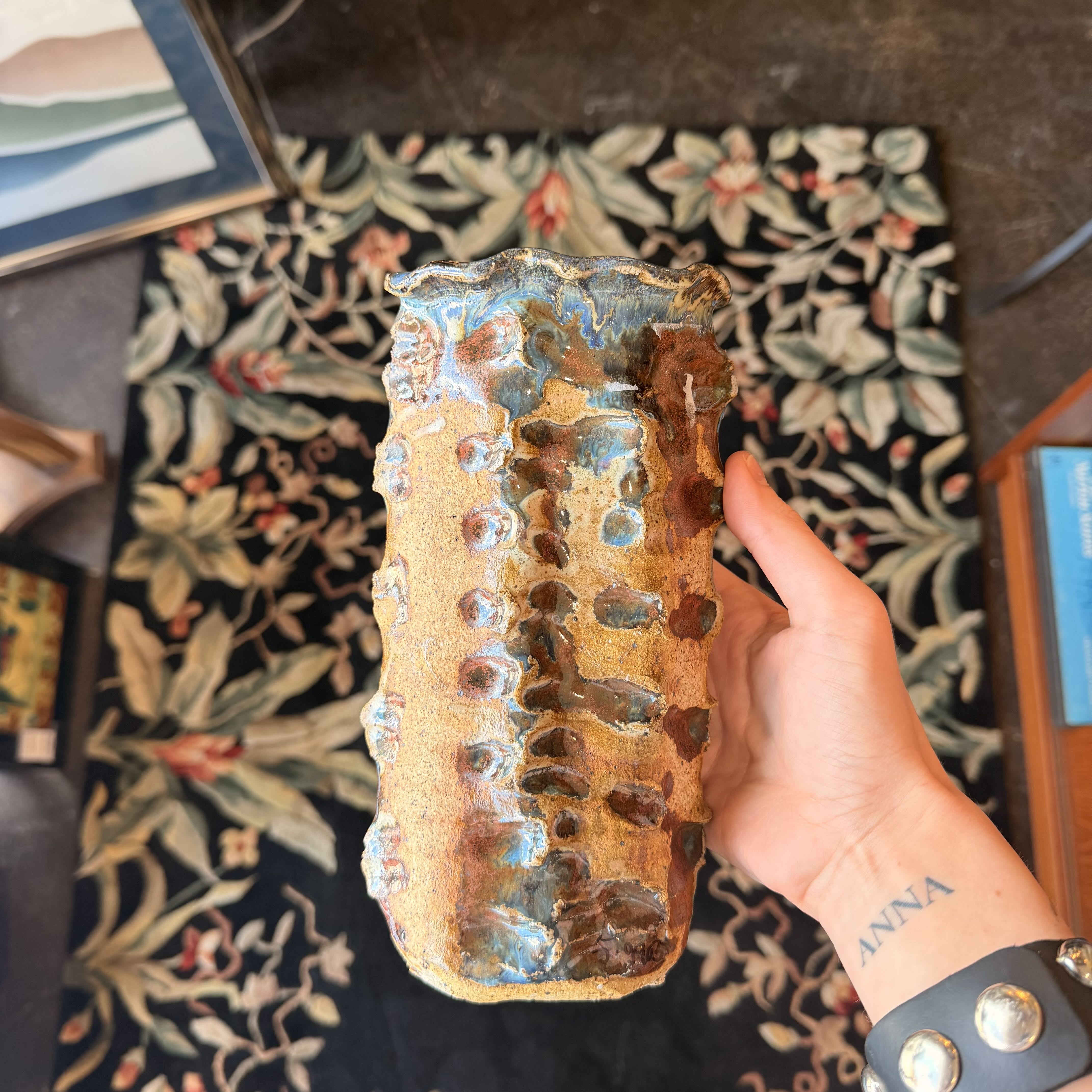 Hand holding a textured ceramic object with a decorative rug in the background