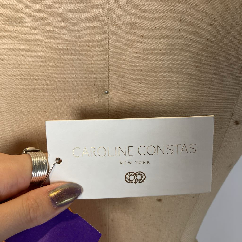 Hand holding a 'Caroline Constas New York' jewelry tag against a beige background
