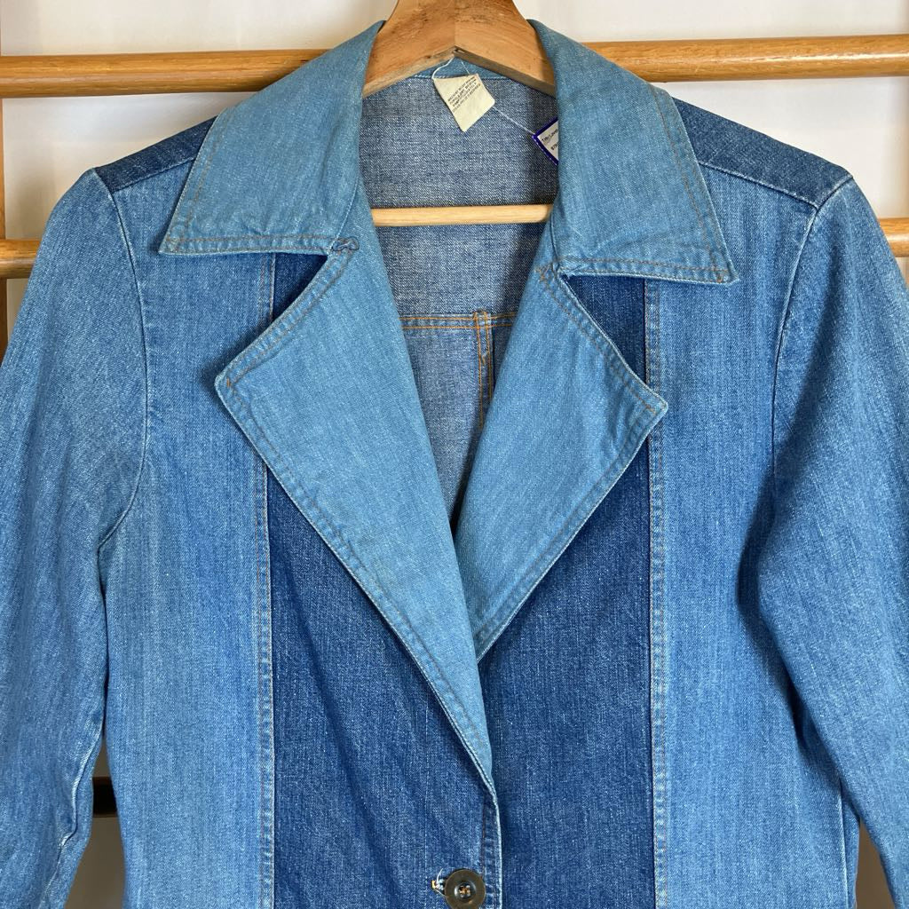 A blue denim jacket with contrasting medium and dark blue denim panels, hanging on a wooden hanger against a light-colored wall.