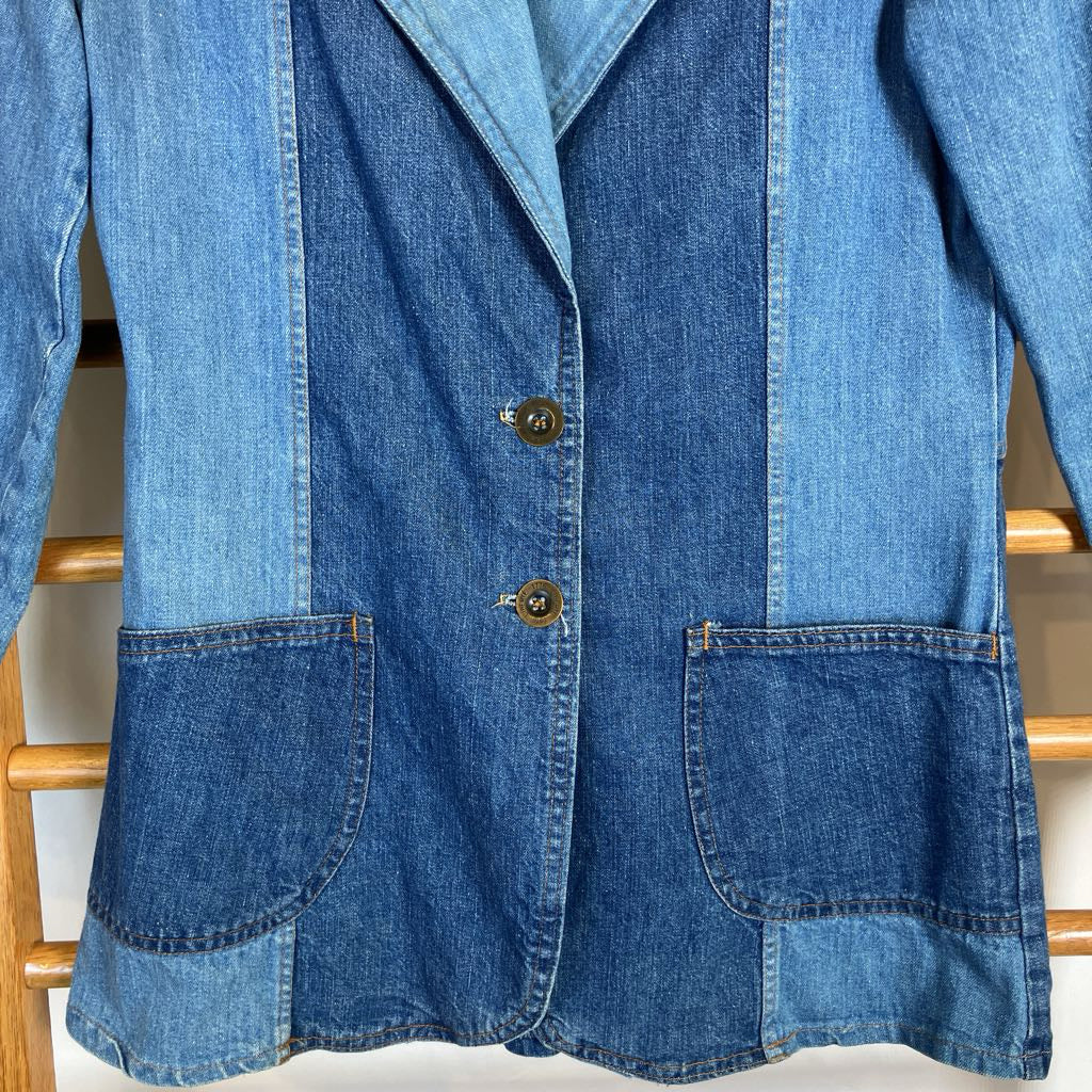 A blue denim patchwork blazer with two front buttons, large front patch pockets, and a notched lapel, shown hanging against a light background.