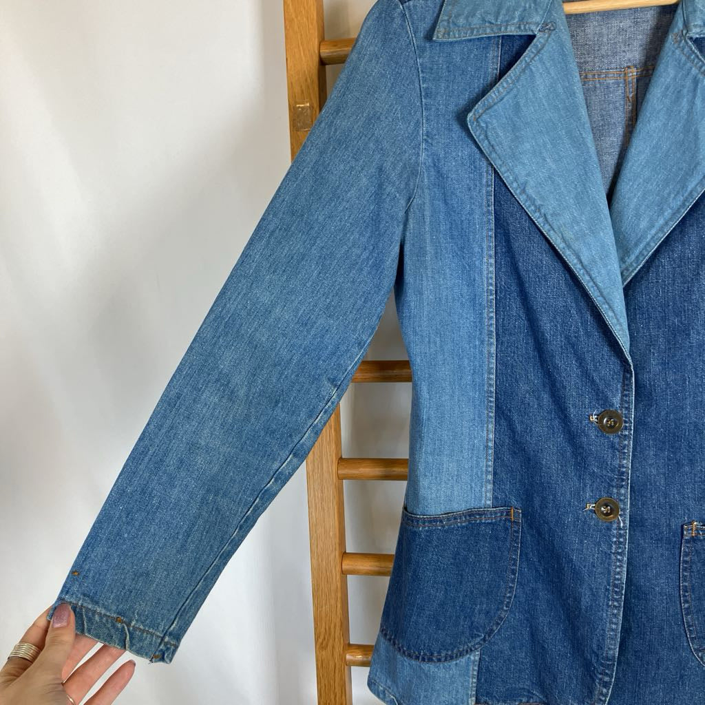A blue denim patchwork blazer jacket hanging on a wooden hanger against a white wall, with a hand pointing to the sleeve.