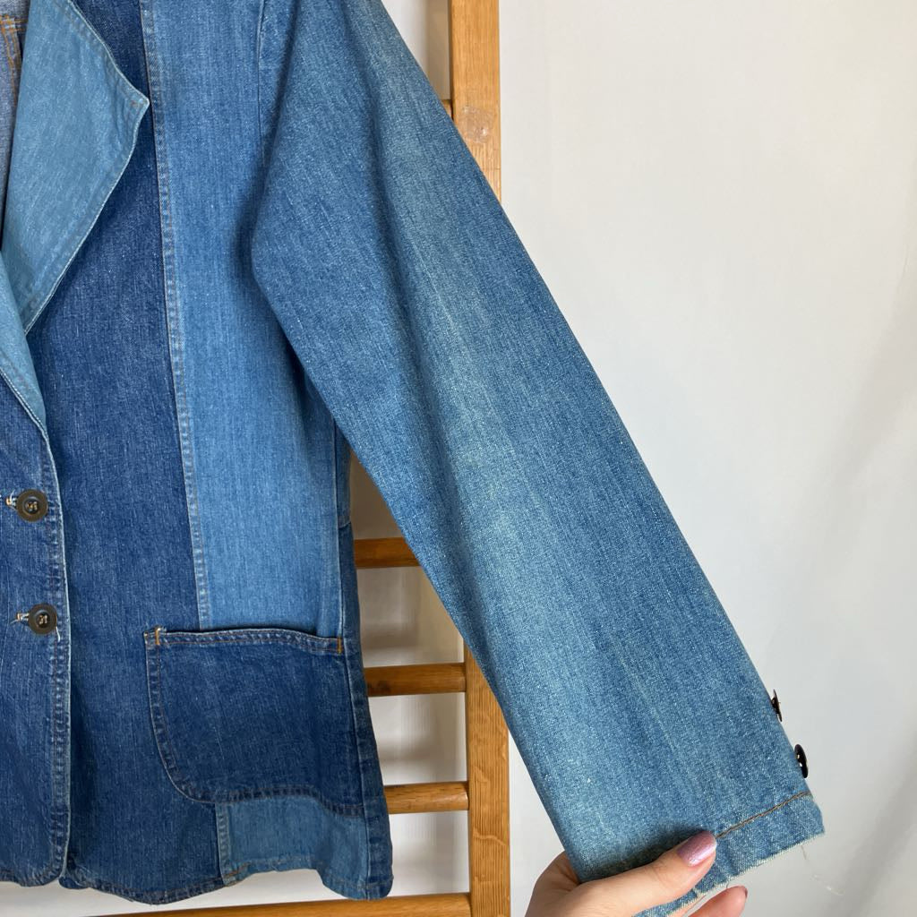 A close-up of a blue denim jacket hanging on a wooden hanger against a plain white wall, with a hand holding the sleeve.