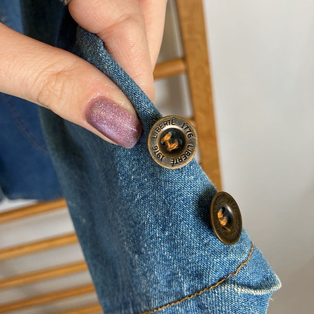 Close-up of a person's hand with glittery mauve nail polish holding a denim jacket sleeve, showing two bronze-toned buttons with 'LIBERTE' engraved on them, against a light background with wooden chair slats.