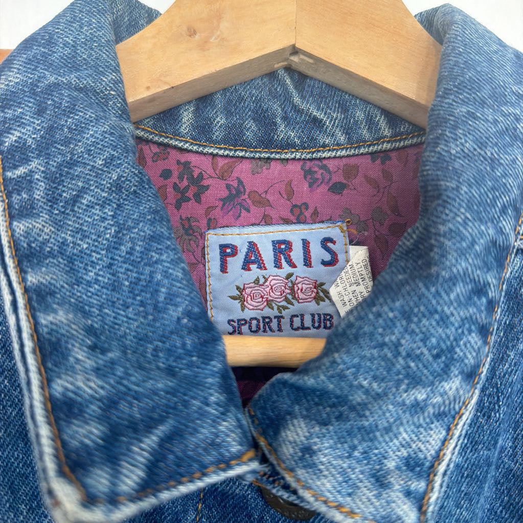 Blue denim jacket with floral lining and 'Paris Sport Club' label on a wooden hanger.