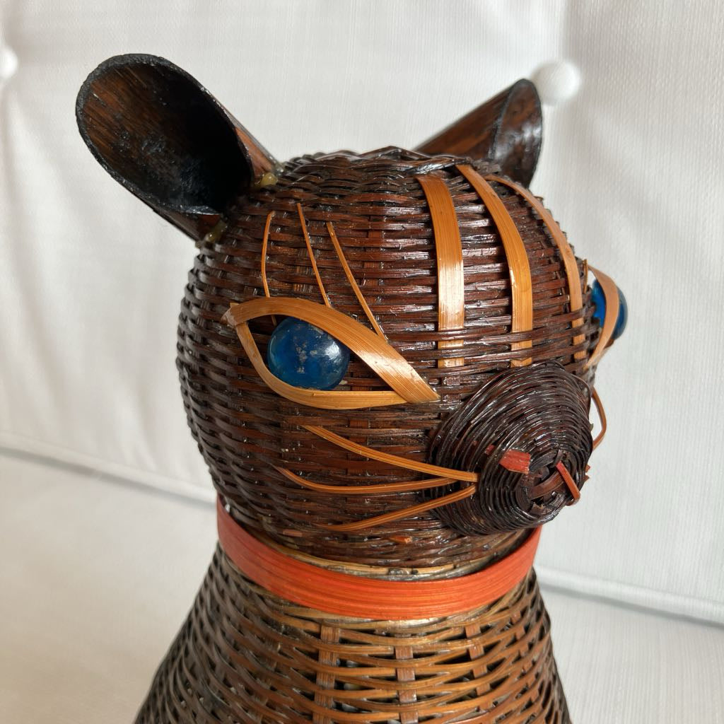 Wicker cat-shaped object with blue glass eyes and orange ribbon on a white background