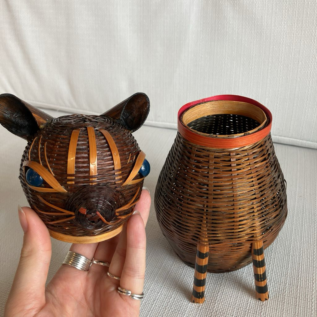 Wicker cat-shaped container held by a hand with rings, next to another similar container on a neutral background.