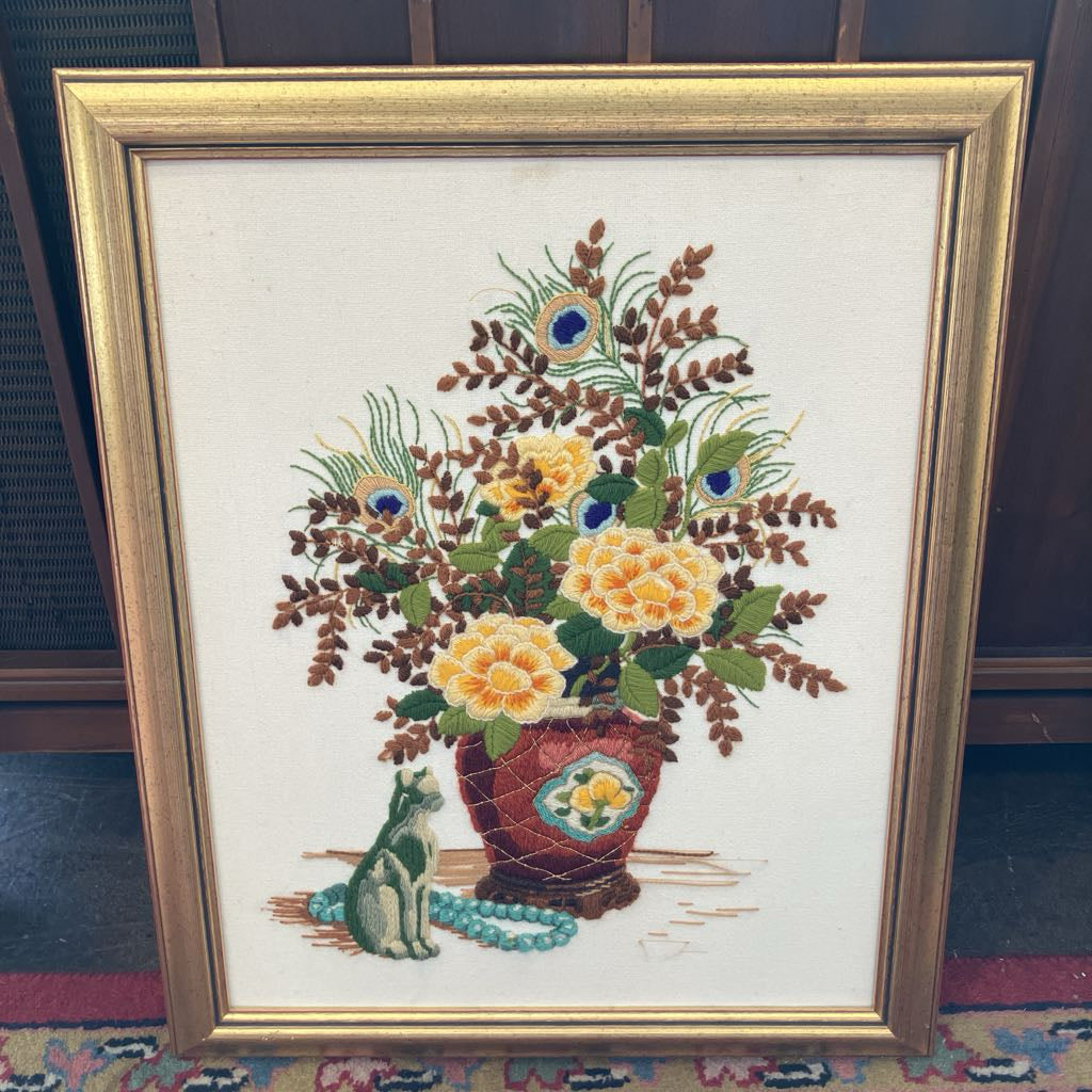 Framed cross-stitched picture of a vase with flowers and a cat on a patterned rug.