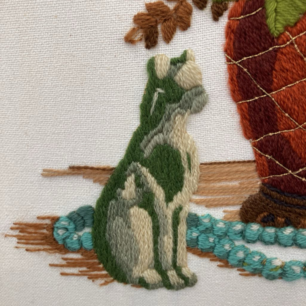 Embroidered design of a cat sitting next to a basket on fabric.