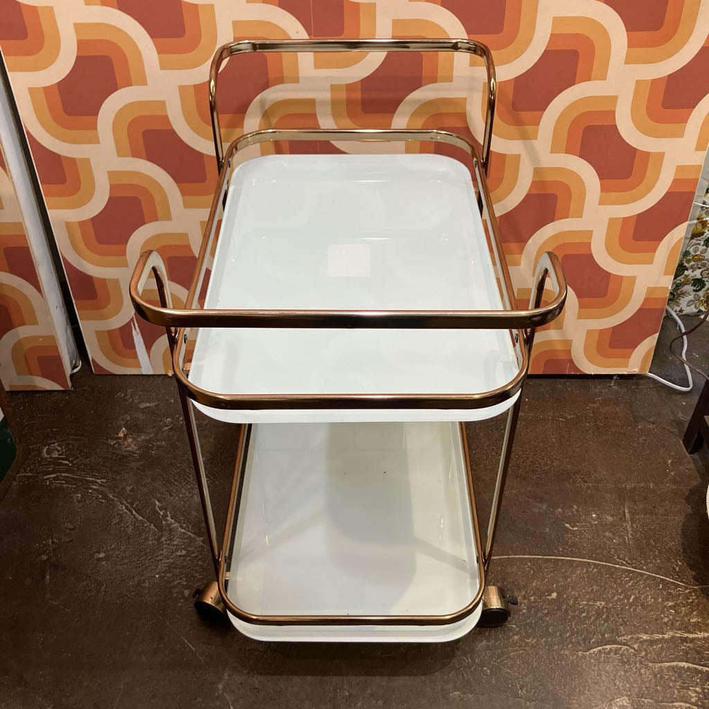Vintage-style bar cart with gold frame and white shelves against a patterned wall.