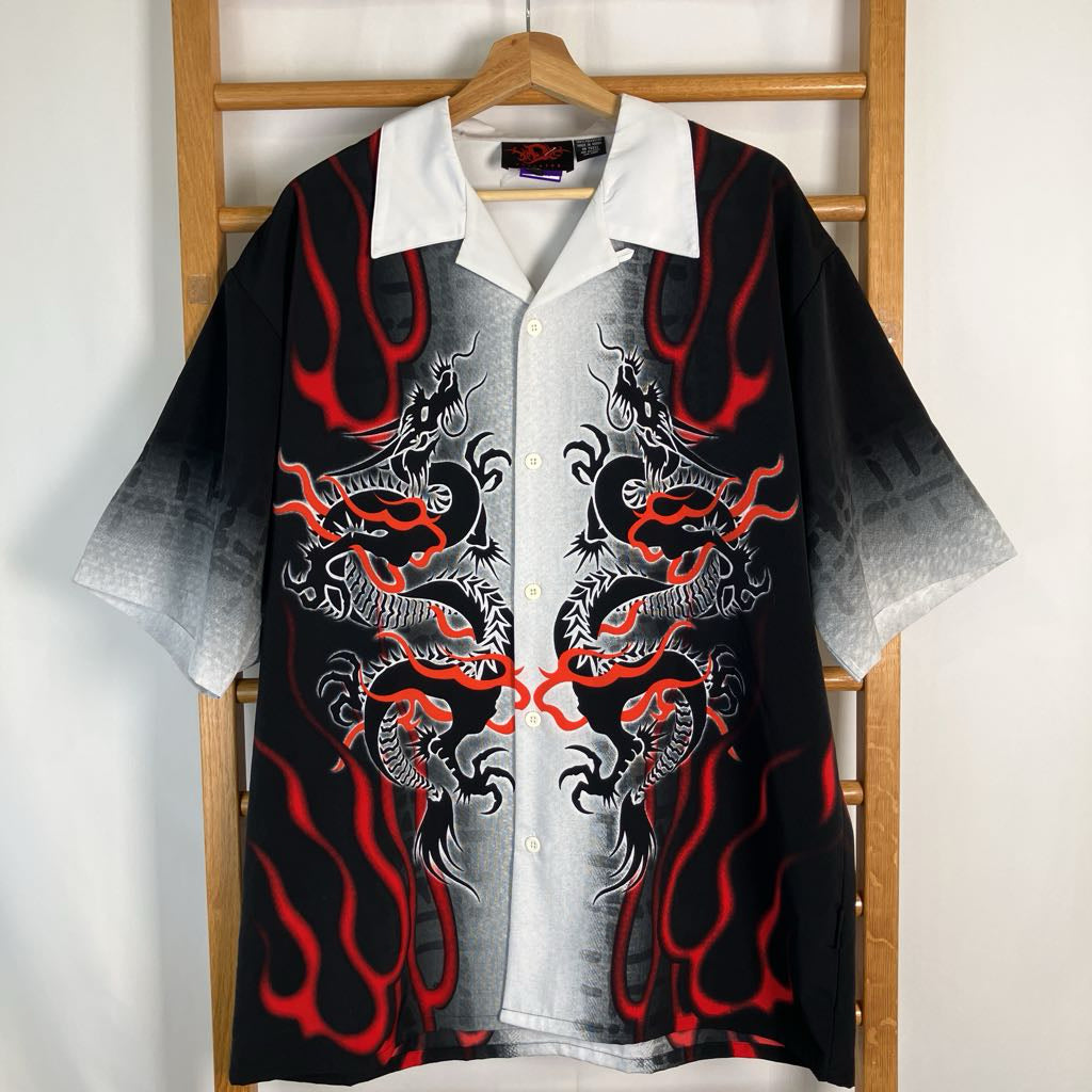A short-sleeve button-up shirt with a black and white gradient body, featuring detailed red and black dragon illustrations and red flame-like patterns, hanging on a wooden hanger against a white wall