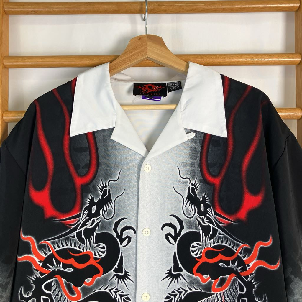 A short-sleeve button-up shirt with a white collar, featuring a black and gray dragon design with red flame accents, hanging on a wooden hanger against a wooden rack.