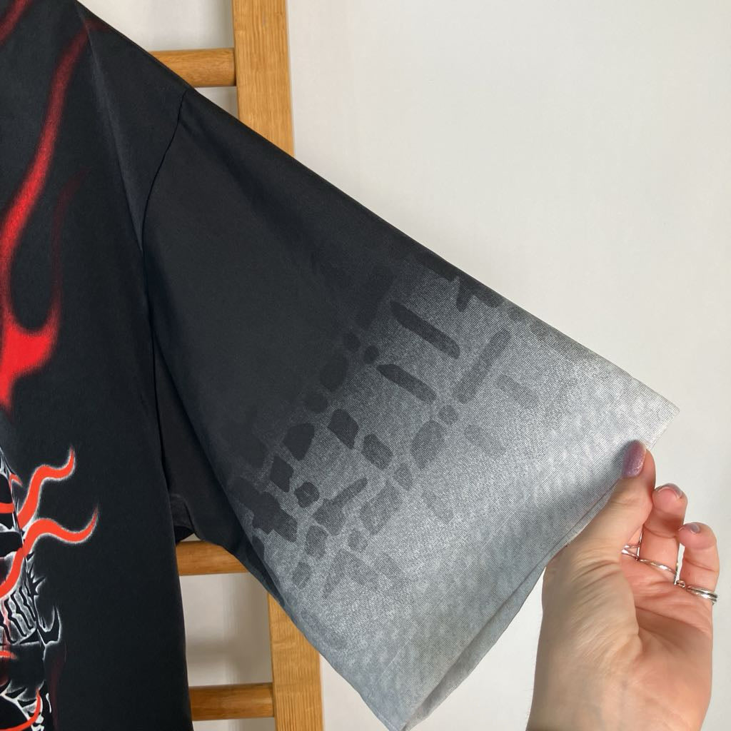 A close-up of a black wide-sleeve shirt with a red flame graphic on the chest and a translucent gray ombré pattern on the sleeve, held by a hand against a white wall.