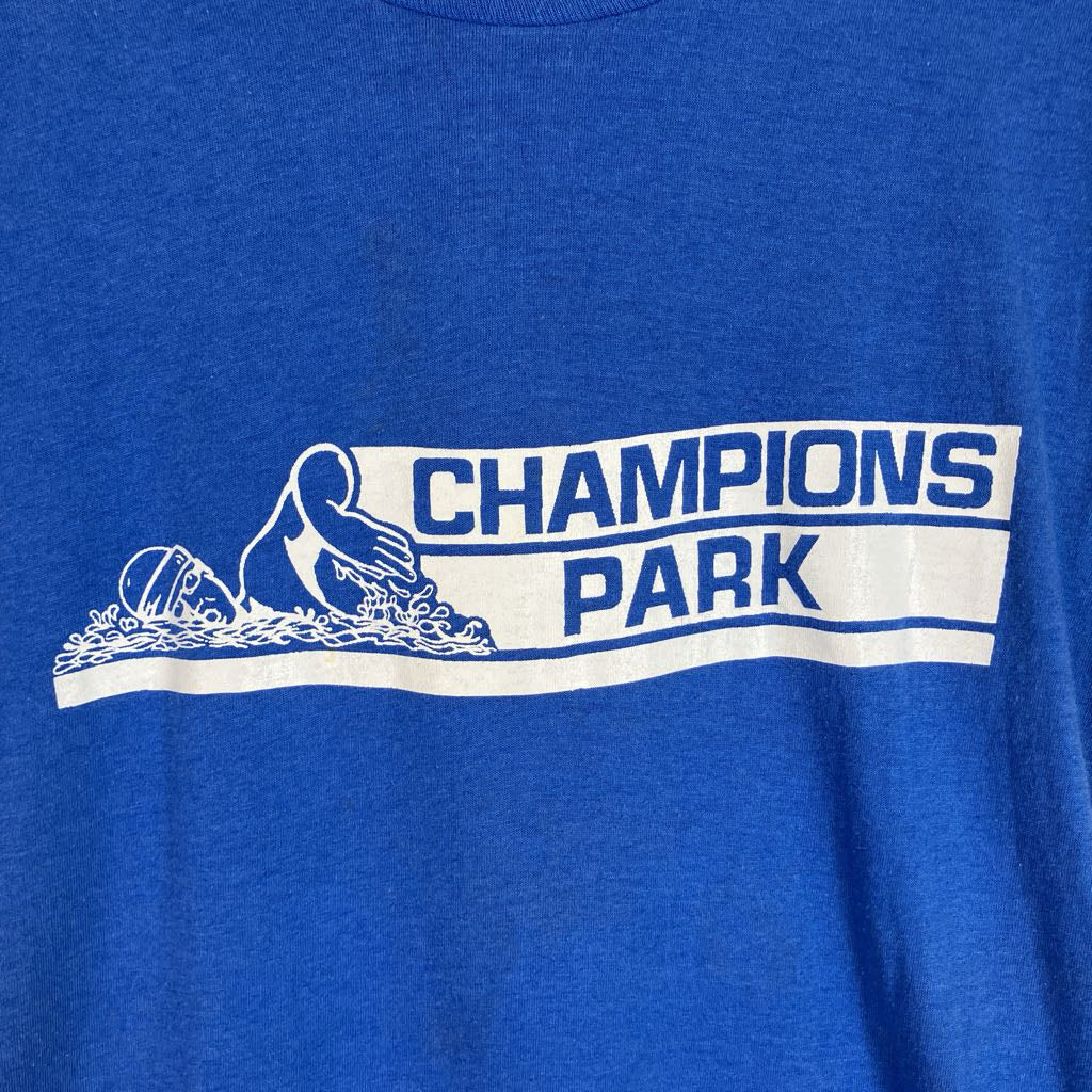 Close-up of a heather royal blue t-shirt featuring a white graphic print that reads 'CHAMPIONS PARK' with a stylized figure and wave illustration.