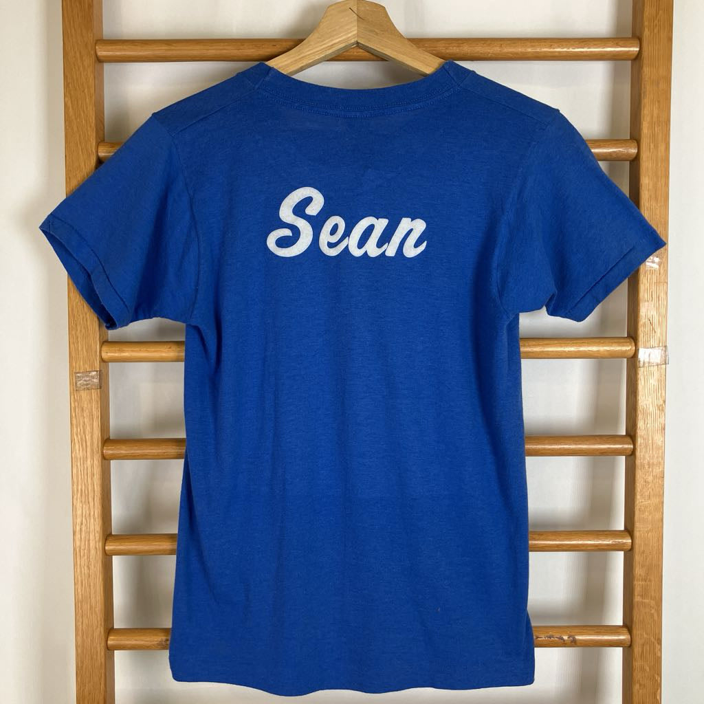 A royal blue short-sleeve t-shirt with the name 'Sean' printed in white script across the chest, hanging on a wooden hanger from a wooden ladder.
