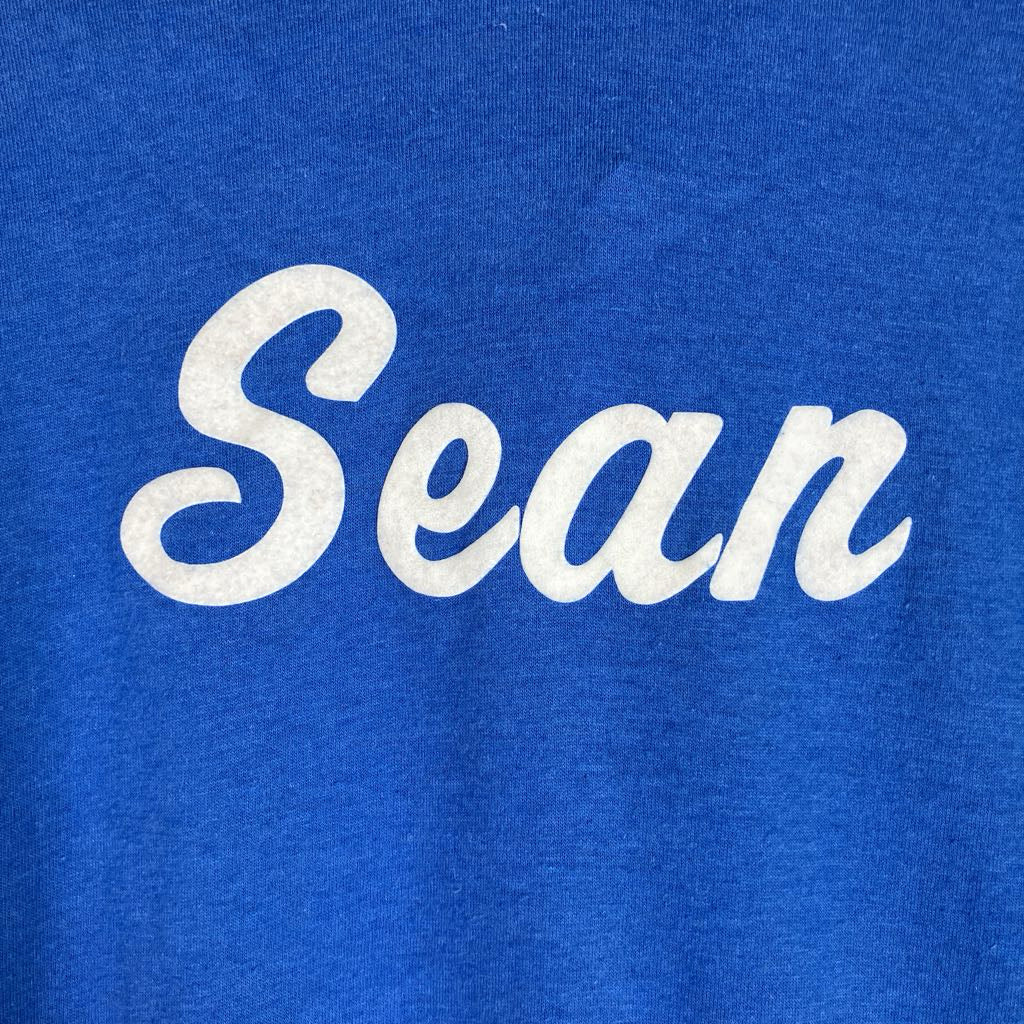 A close-up of a royal blue heathered t-shirt featuring the name 'Sean' printed across the chest in a white cursive script font.