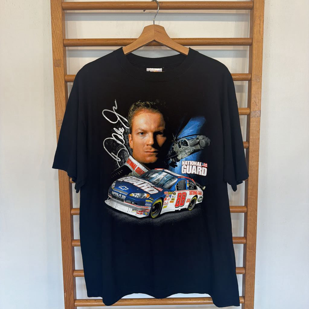 Black t-shirt with a graphic of a race car driver and car on a hanger against a wooden ladder background.