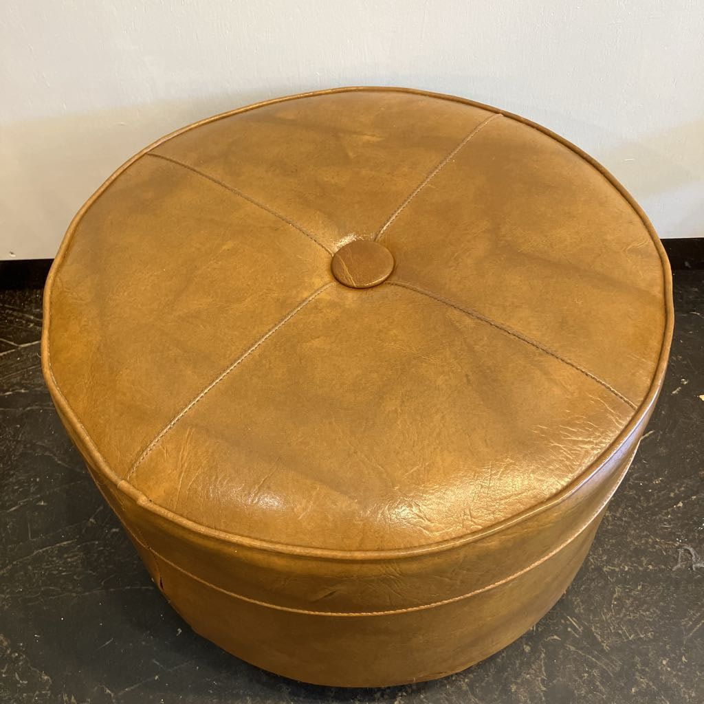 Round brown leather ottoman on a dark floor with a white wall background