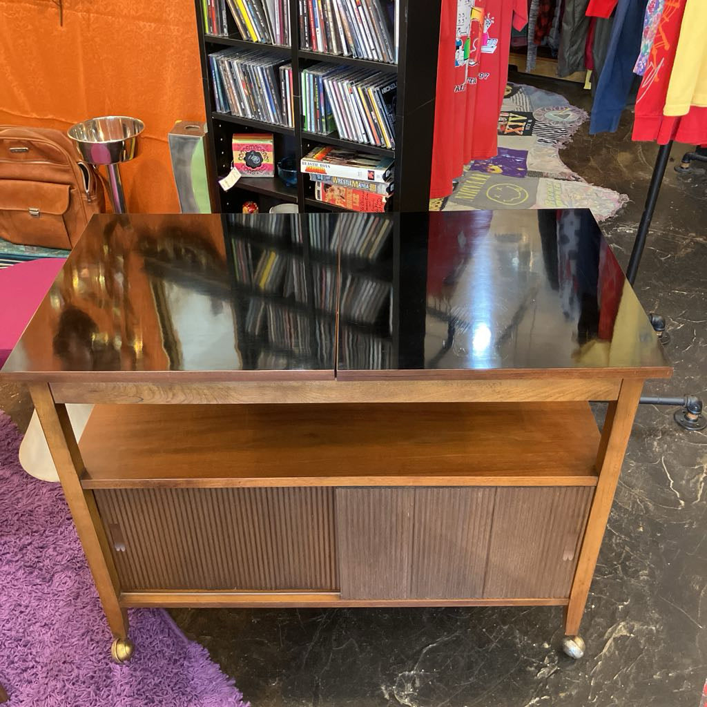 Wooden bar cart with glass top in a room with books and clothing.