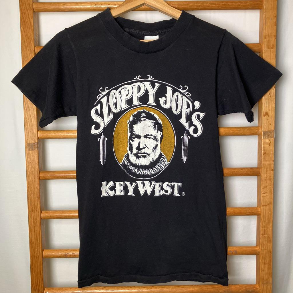 A black vintage graphic t-shirt hanging on a wooden hanger against a wooden ladder background. The shirt features a distressed white graphic reading 'SLOPPY JOE'S KEYWEST' with a circular portrait of a bearded man in the center.