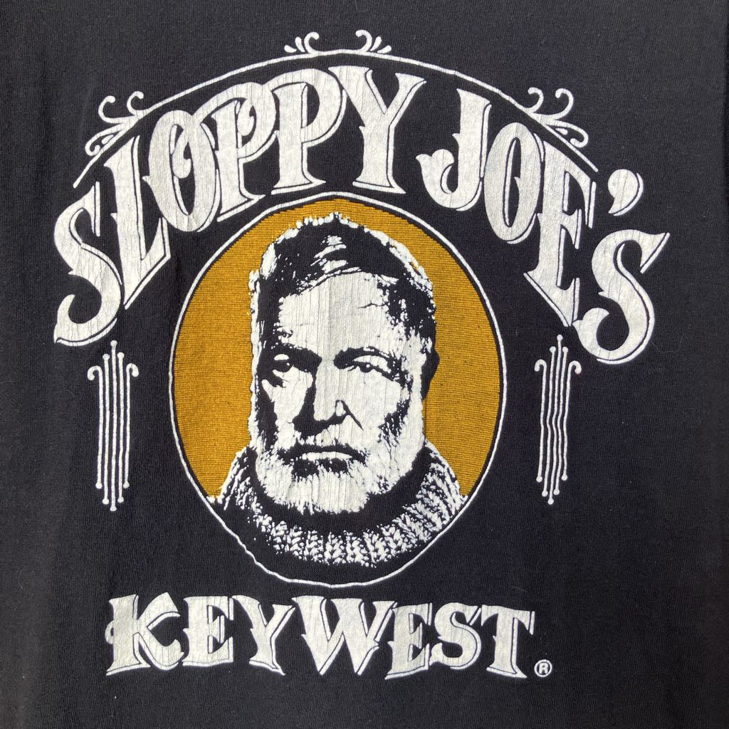 Close-up of a black graphic print featuring the words 'SLOPPY JOE'S KEY WEST' in white decorative lettering around a circular portrait illustration.