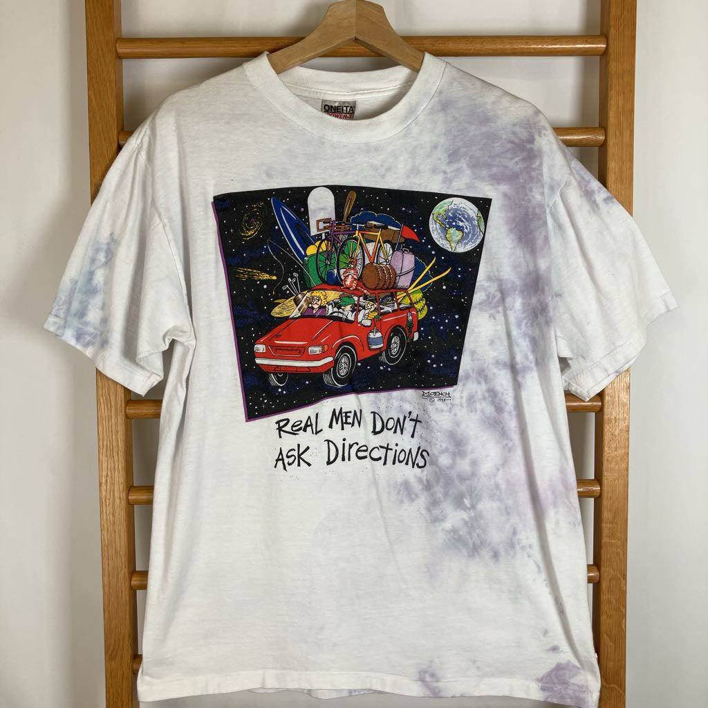 A white tie-dye t-shirt with a colorful graphic of a red car carrying a surfboard, a helmet, and a globe, with the text 'REAL MEN DON'T ASK DIRECTIONS' printed below it. The shirt is hanging on a wooden hanger on a wooden chair.