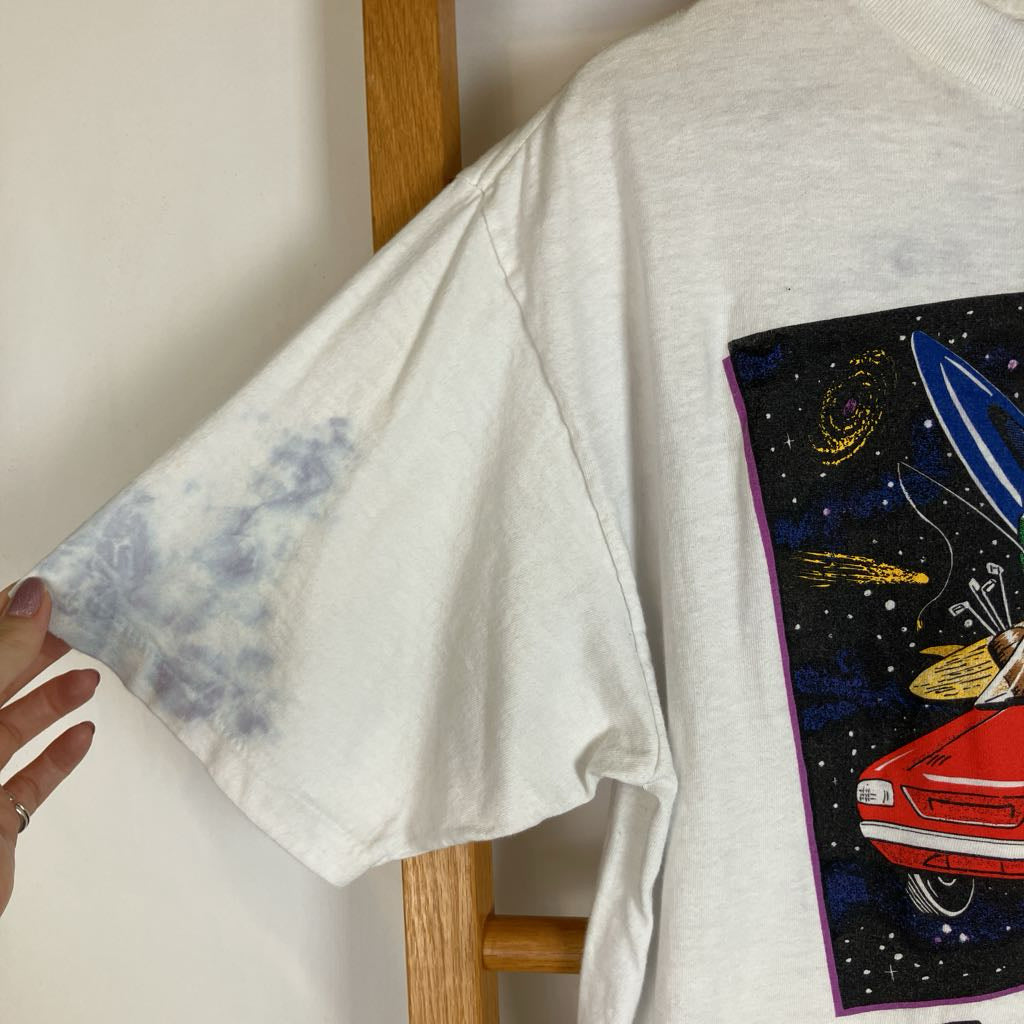 A white oversized graphic T-shirt hanging on a wooden hanger, featuring a large multicolor space-themed print on the front with a red car and a blue sleeve with a gray tie-dye wash. A hand is holding the sleeve.