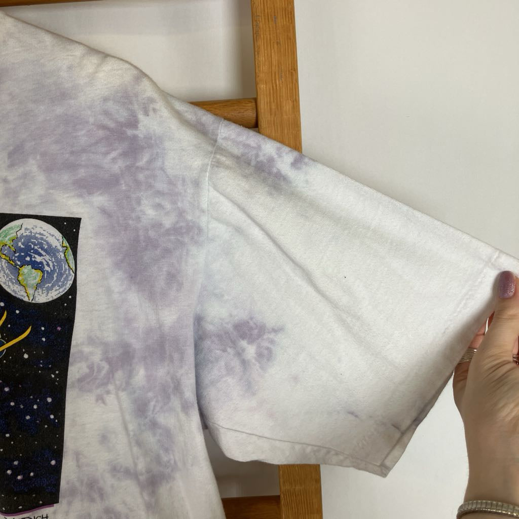 A white short-sleeve t-shirt with a light purple tie-dye pattern and a graphic print of the Earth on the chest, held up by a wooden hanger with a hand visible on the right.