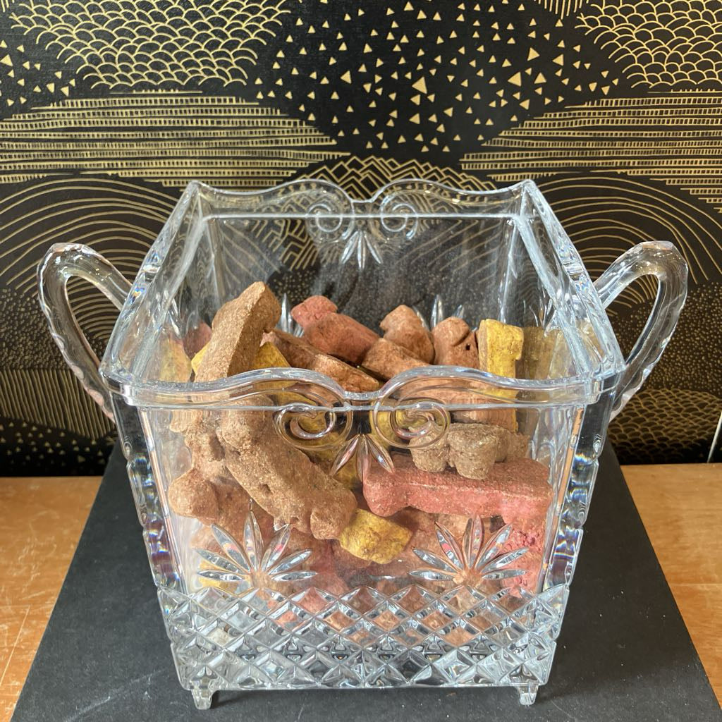 Clear glass candy dish filled with dog treats on a black surface with a decorative gold background.