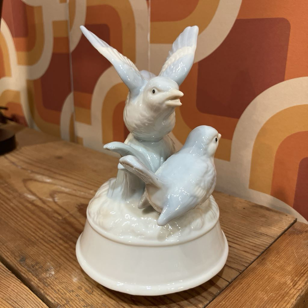 White ceramic sculpture of two birds on a wooden surface with a patterned wall in the background