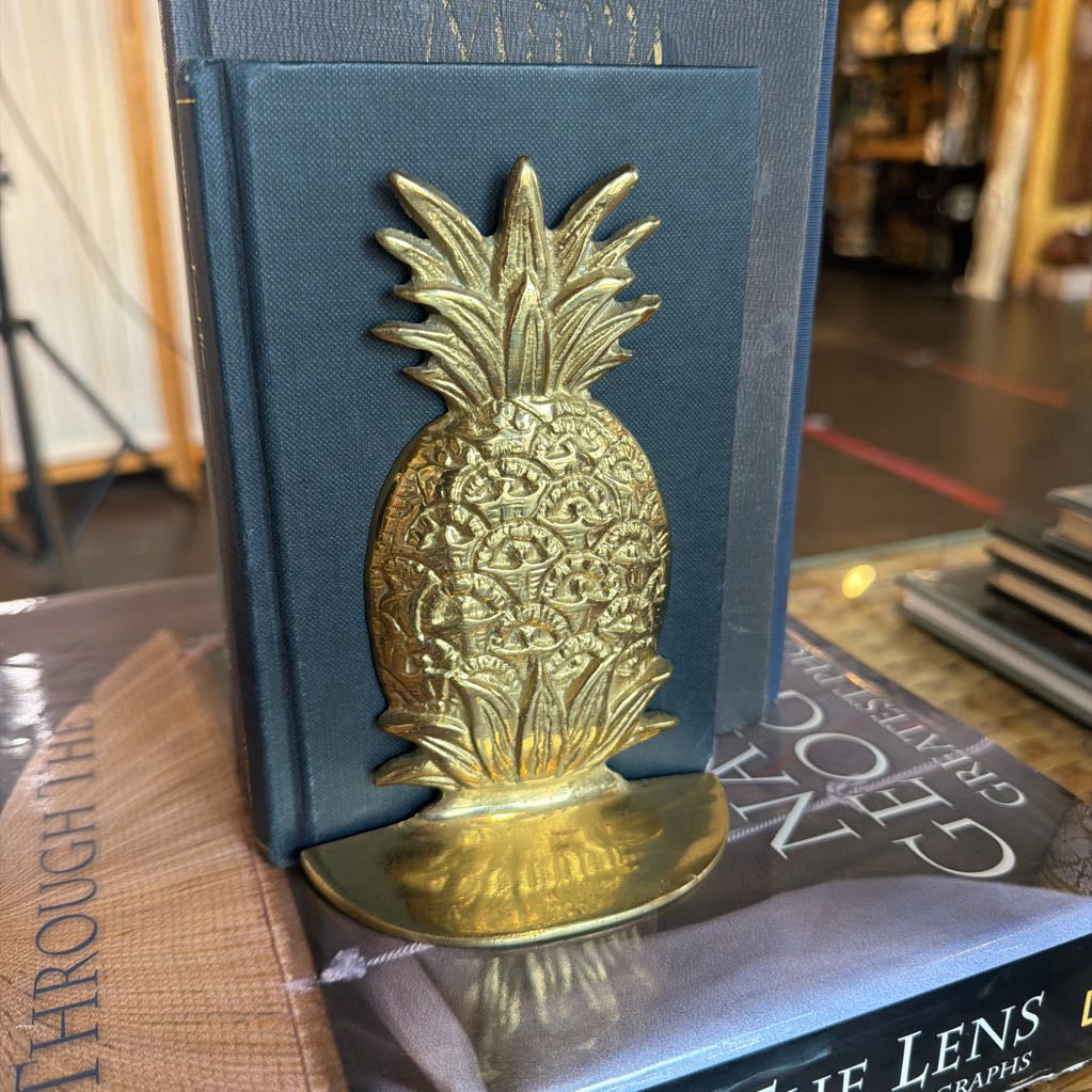 A brass pineapple bookend holding a dark blue hardcover book, with other books visible in a blurred indoor setting.