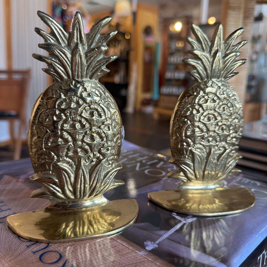 Two ornate pineapple-shaped brass decorative sculptures with intricate floral engravings and round bases, displayed on a reflective surface in a softly blurred indoor setting.