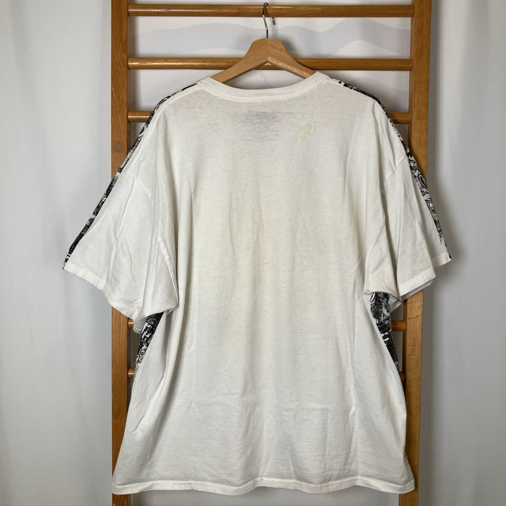 White t-shirt with black patterns on sleeves hanging on a wooden rack against a white wall.