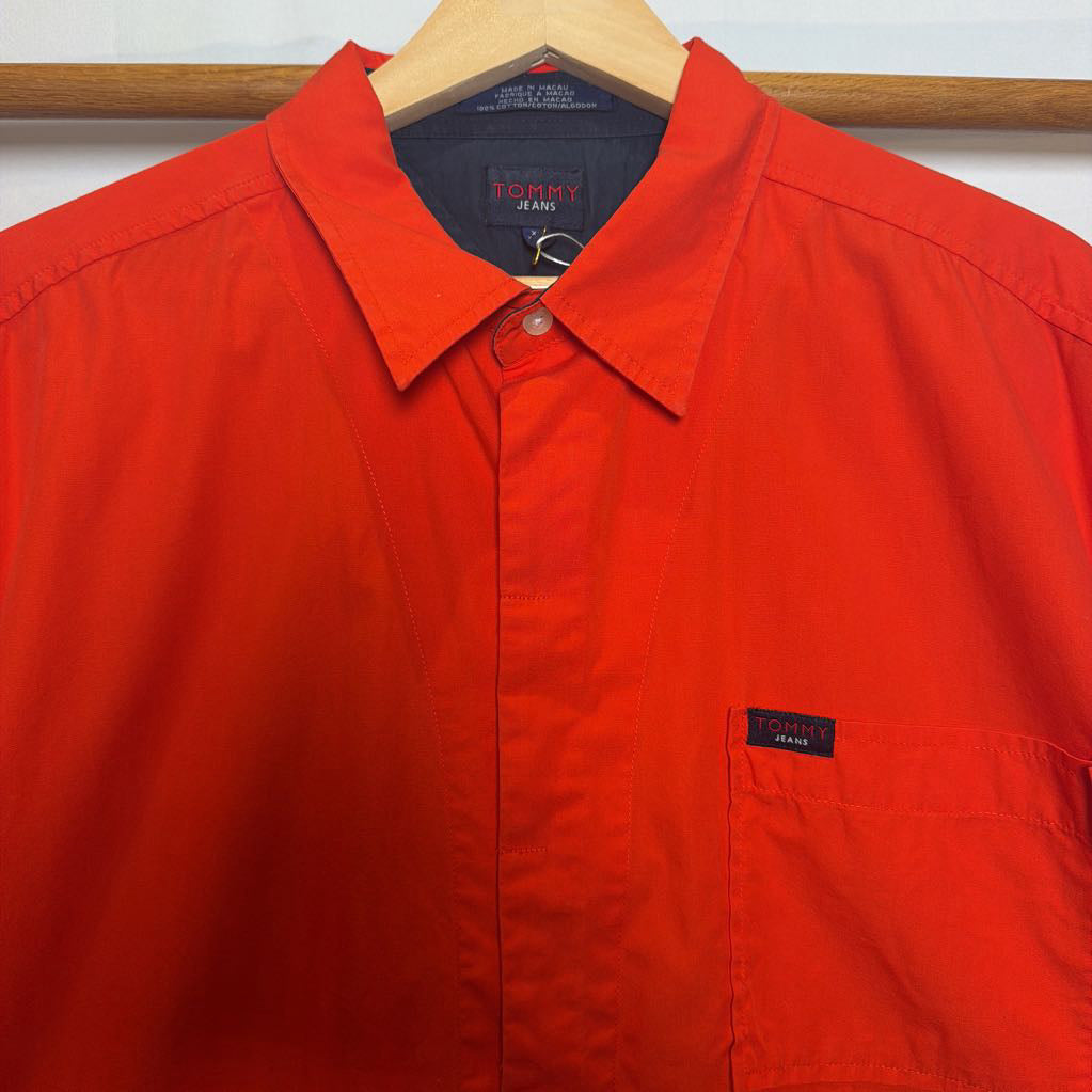 A bright red Tommy Jeans button-up shirt hanging on a wooden hanger against a white wall.