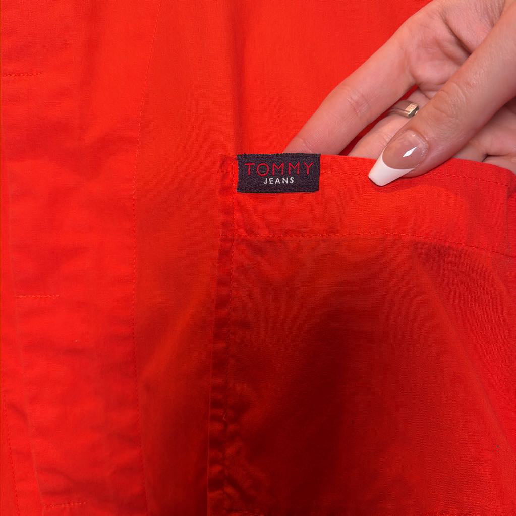A close-up of a bright red Tommy Jeans garment with a hand pointing to the brand label on the pocket.