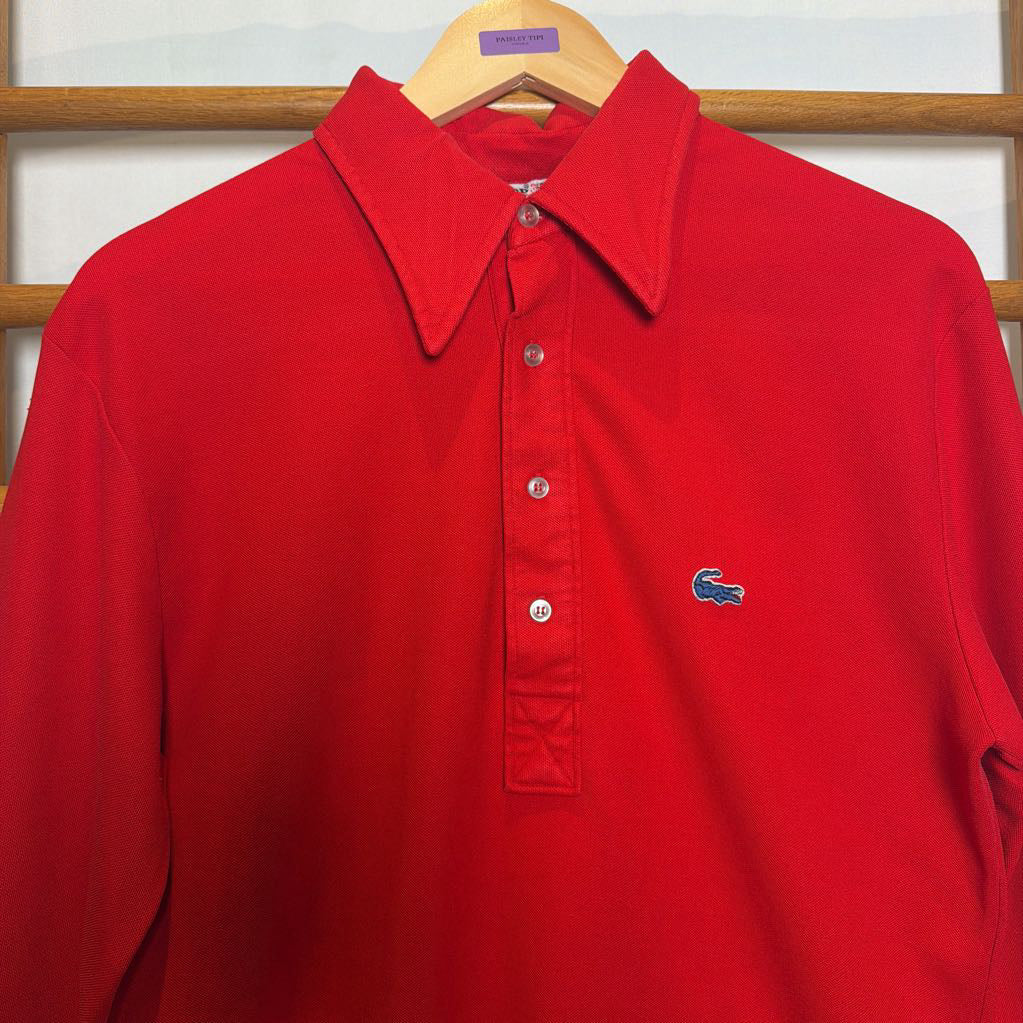 A bright red Lacoste long-sleeve polo shirt hanging on a wooden hanger against a light-colored paneled background. The shirt features a classic pointed collar, a three-button placket, and the signature blue crocodile logo embroidered on the left chest.