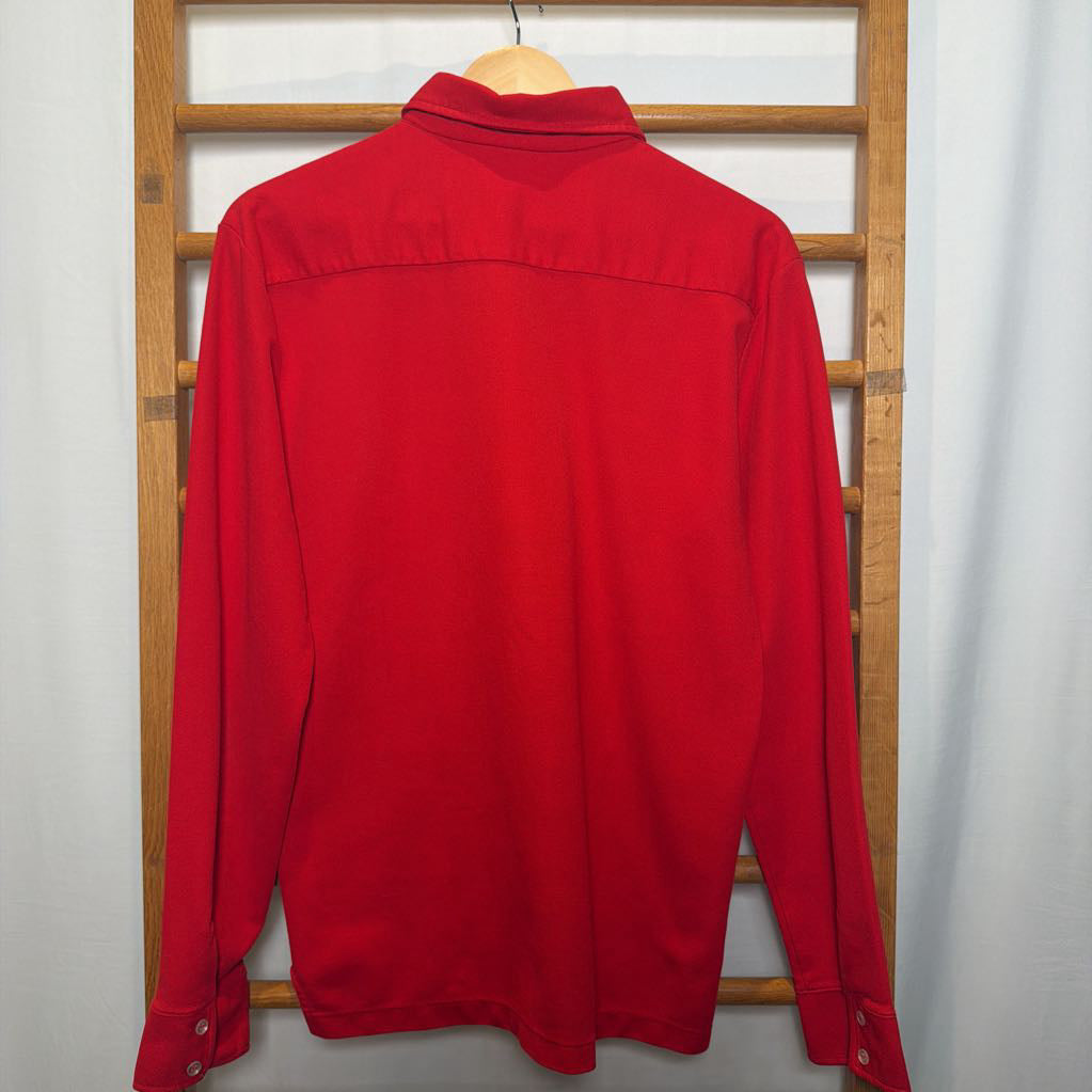 A bright red long-sleeve collared shirt hanging on a wooden hanger against a white curtain backdrop with a wooden ladder visible behind it.