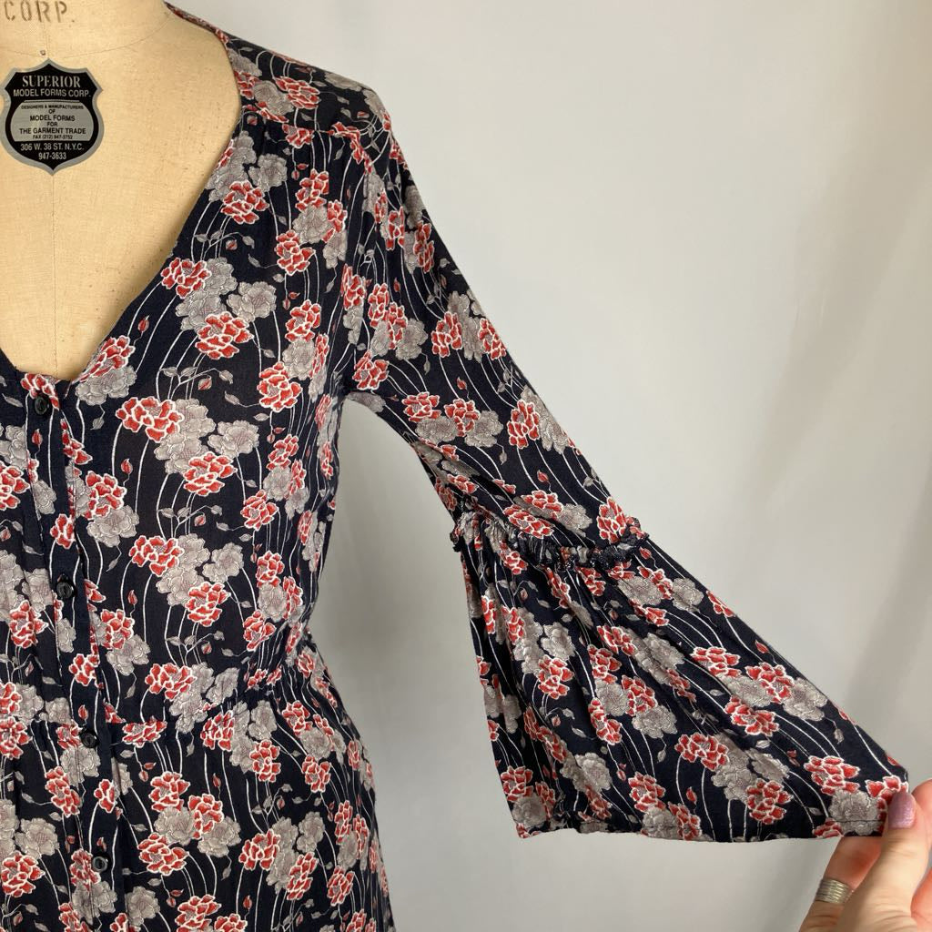 A close-up of a sleeveless V-neck dress on a mannequin, featuring a navy blue base with a red and gray floral pattern. The dress has a gathered waist and a flared bell sleeve, with a hand holding out the sleeve to show its width.