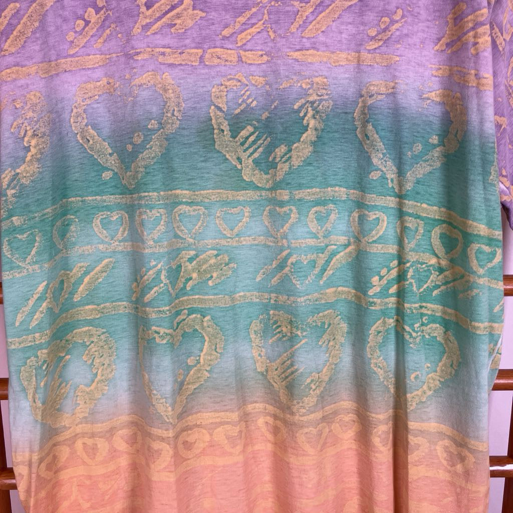 A close-up of a fabric garment featuring a vertical gradient design transitioning from lavender purple at the top, through teal in the middle, to peach-pink at the bottom, decorated with metallic gold heart and tribal-style patterns.