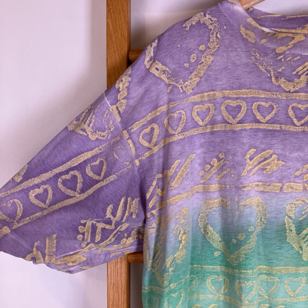 A close-up of a short-sleeve t-shirt featuring a purple-to-teal gradient with distressed gold heart and tribal-style patterns, hanging against a light wall and wooden hanger.