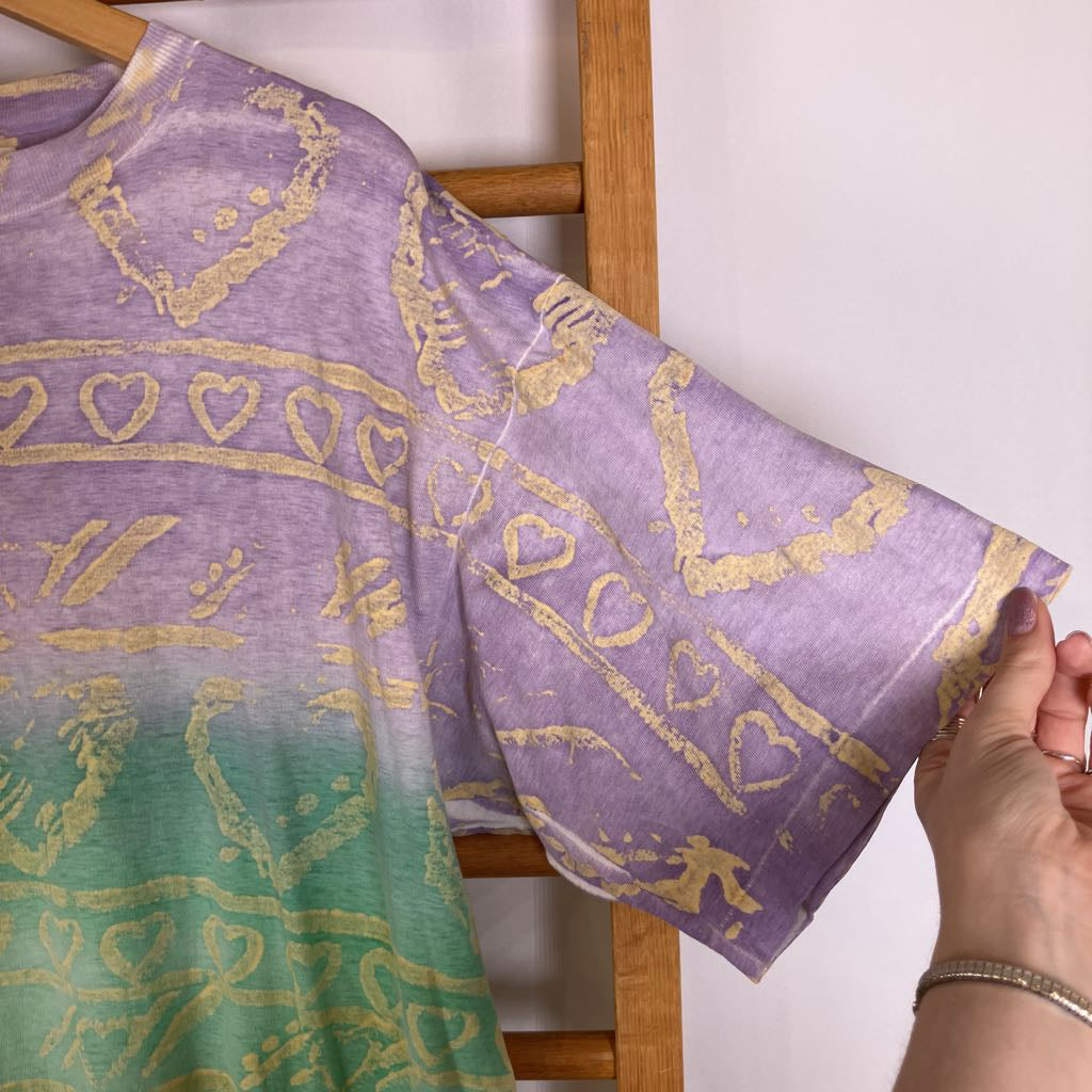 A lavender and green gradient short-sleeve t-shirt with gold metallic foil patterns, held up by a wooden hanger with a person's hand visible on the right side.