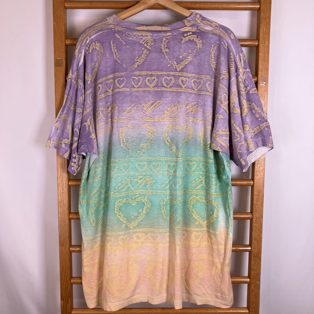 A short-sleeve t-shirt with a purple-to-teal-to-cream gradient and gold heart patterns, hanging on a wooden hanger against a white wall.