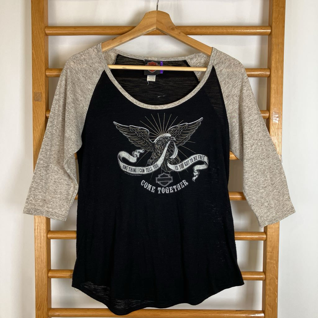 A women's raglan baseball t-shirt hanging on a wooden hanger against a wooden ladder. The shirt has a black body with heather gray three-quarter sleeves and a distressed white graphic on the front featuring an angel, a ribbon, and the text 'COME TOGETHER' with 'WE WILL SEE' and 'IN TIME TO COME'.