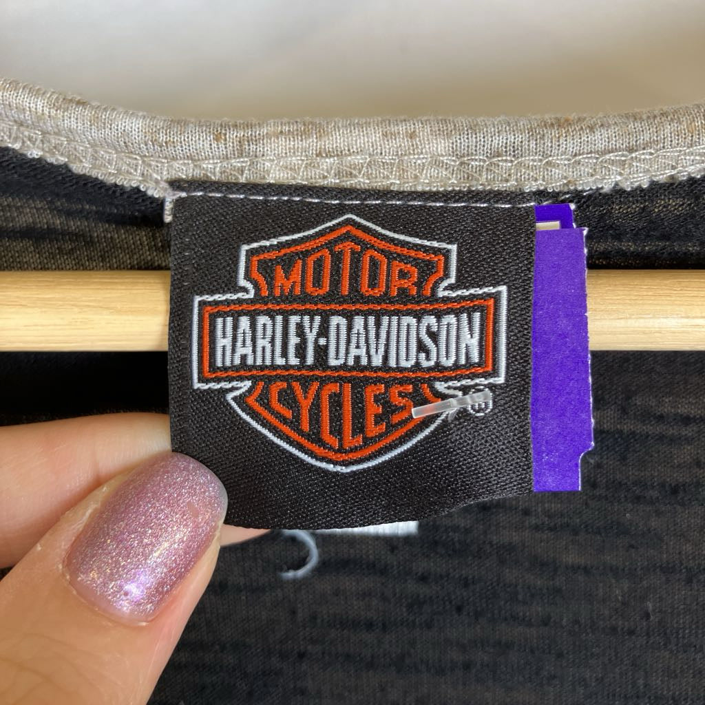 A close-up of a black fabric tag with the Harley-Davidson Motor Cycles logo, held by a hand with glittery pink fingernails. The tag is sewn onto a dark gray garment with a light wood dowel behind it.