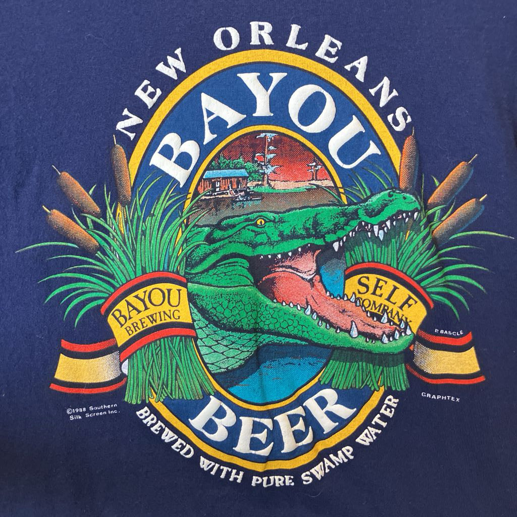 A close-up of a navy blue graphic print featuring a green alligator, cypress trees, and the text 'New Orleans Bayou Beer' with 'Self Owned' on a banner.