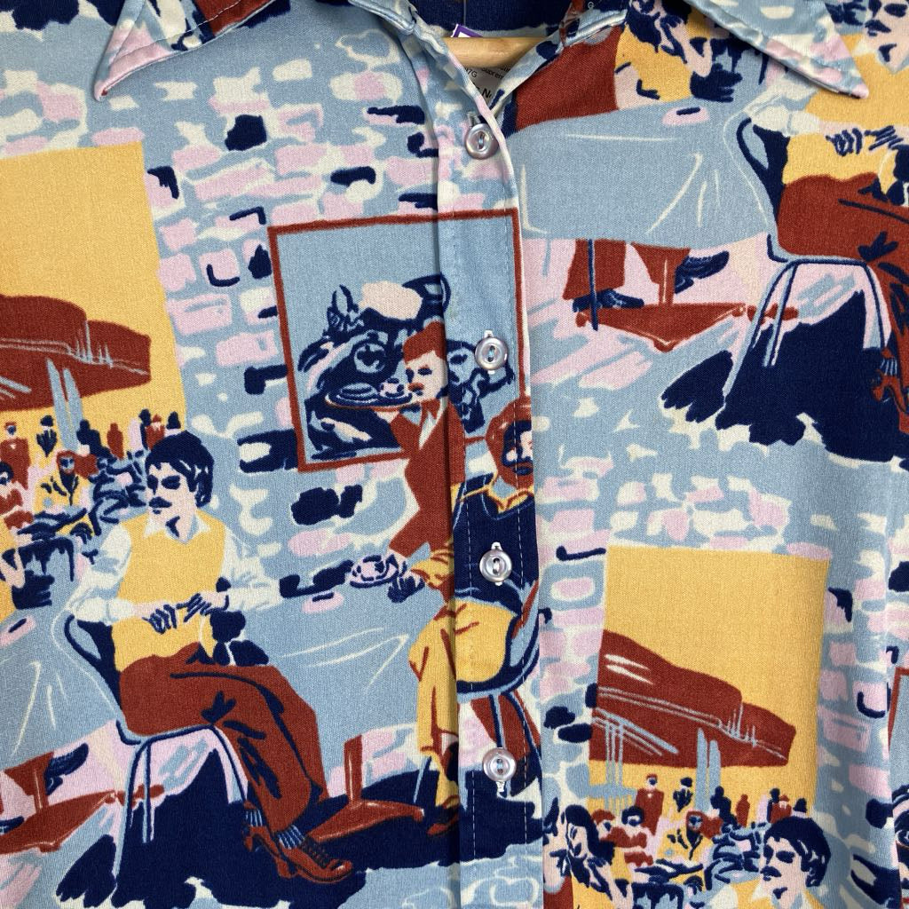 A close-up of a vintage button-up shirt featuring a bold, multicolor graphic print with illustrations of figures, chairs, and abstract shapes in light blue, yellow, red, and navy.