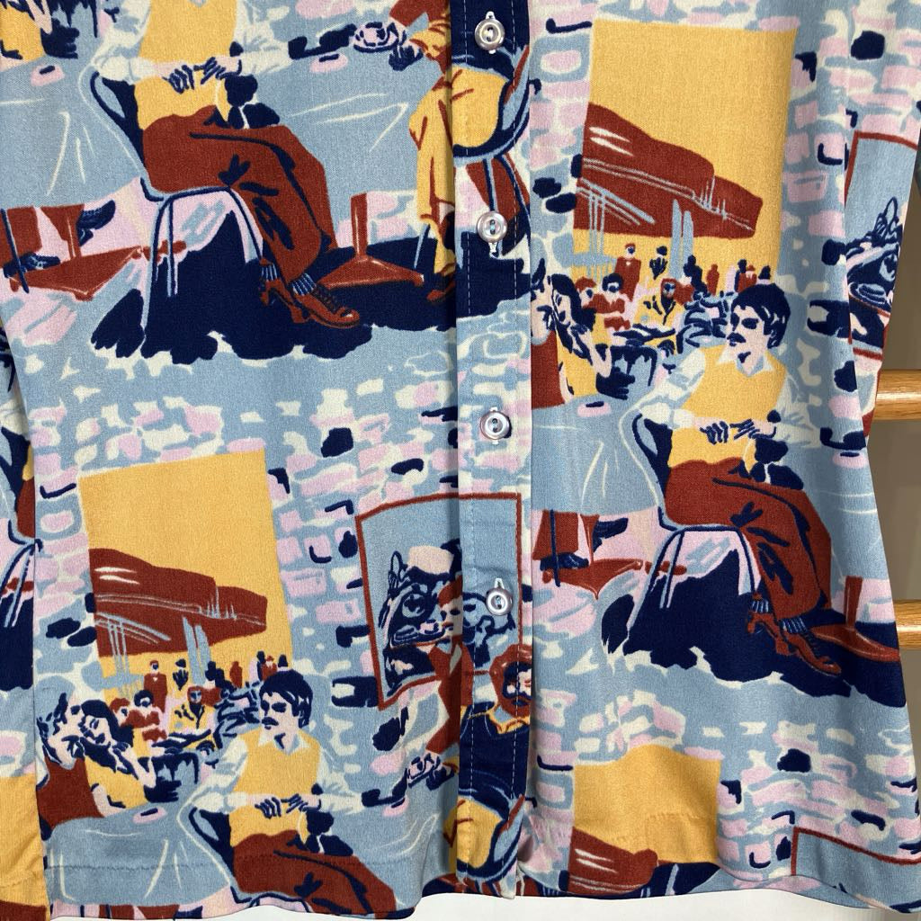 A close-up of a button-front shirt or jacket fabric with a busy illustrated print. The design shows figures, chairs, and architectural shapes in light blue, mustard yellow, navy, brown, pink, and white.