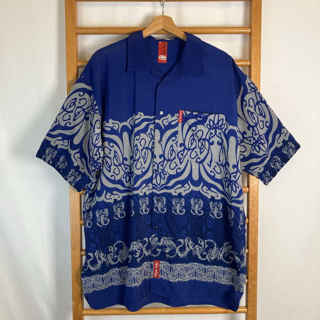 A blue short-sleeve button-up shirt with a white and gray traditional batik-style pattern, hanging on a wooden hanger on a wooden clothes rack against a white wall.