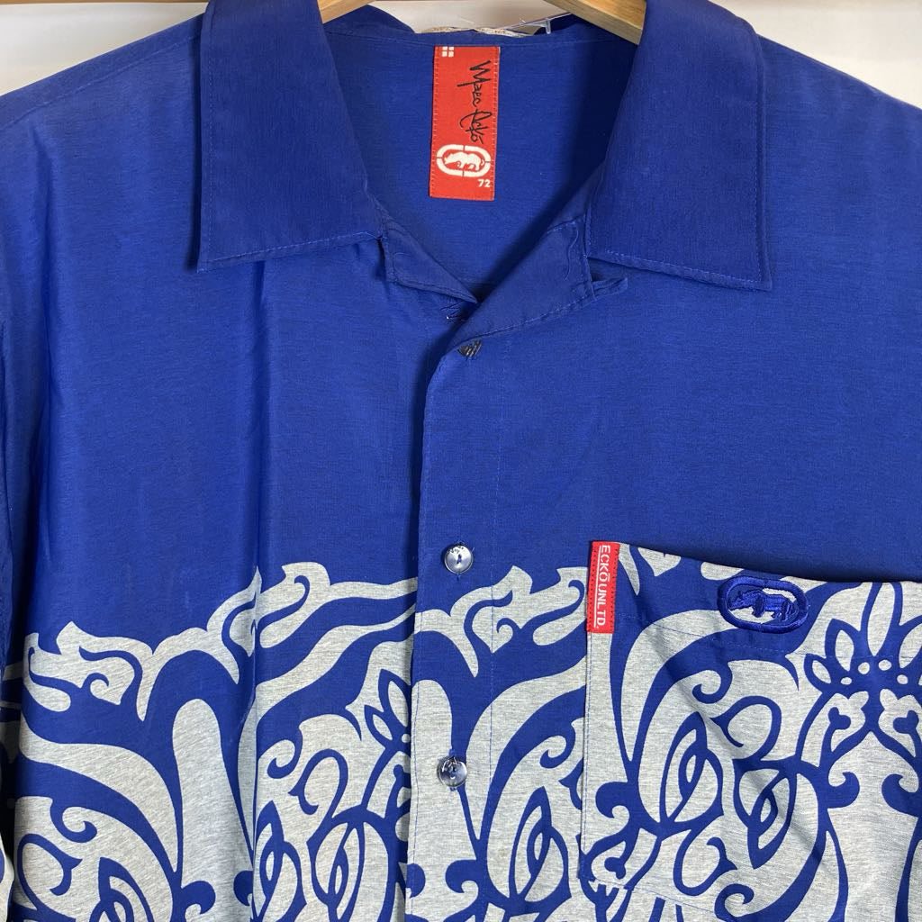 A men's short-sleeve camp collar button-up shirt in royal blue, featuring a light gray heathered lower panel with a blue traditional wave pattern and a blue embroidered logo on the chest pocket. The shirt is shown hanging on a wooden hanger against a white background.