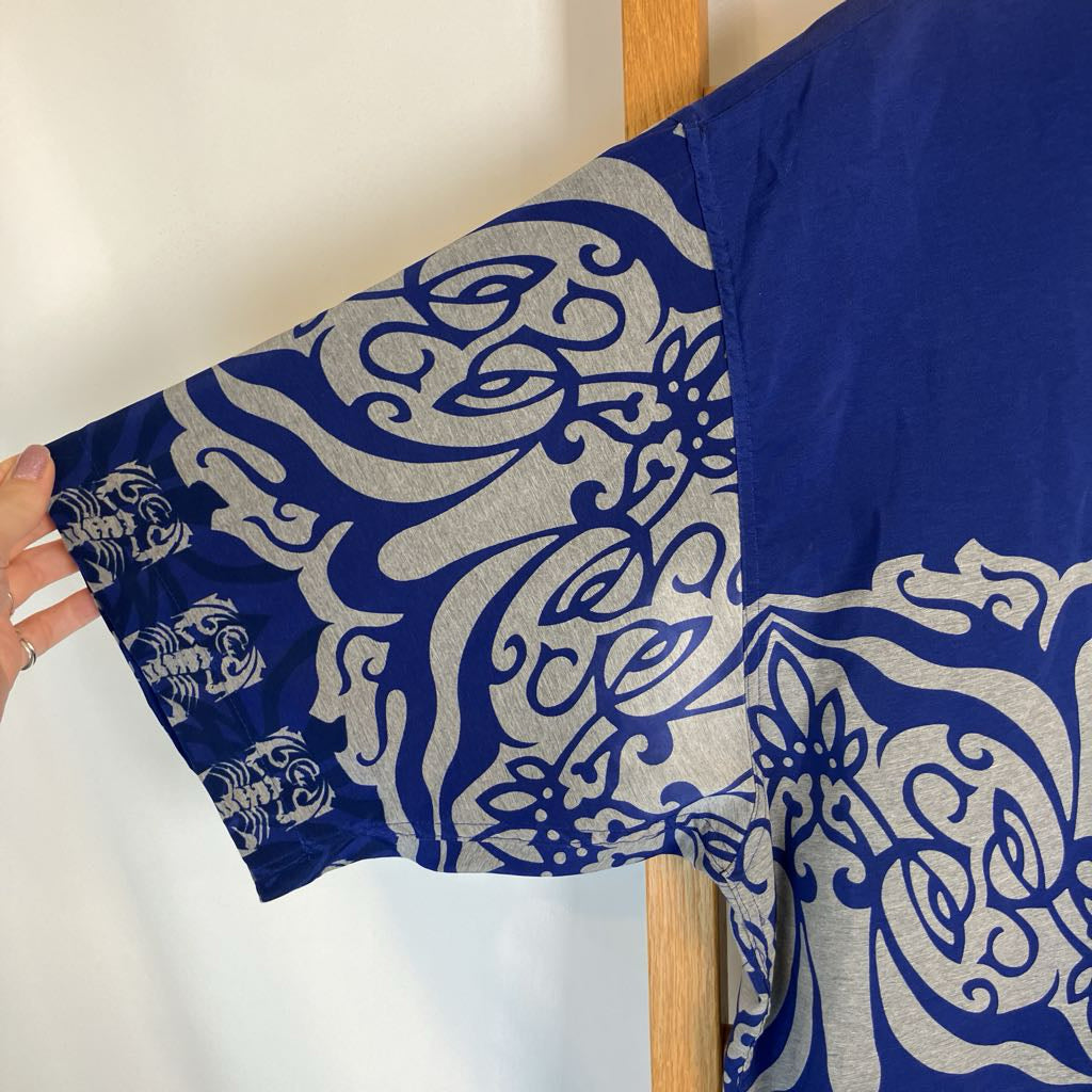 A close-up of a royal blue garment with a silver-gray ornate scroll and floral pattern, held by a hand on the left side against a plain light background.
